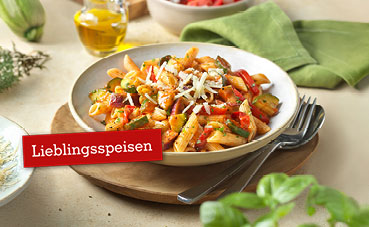 Gedeckter Tisch mit einer Portion Penne mit mediterraner Gemüsesauce aus Tomaten, Zucchini, Melanzani, Zwiebel und Basilikum.