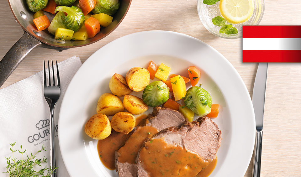Ein Teller mit einem klassischen Schweinsbraten mit Bratkartoffeln und Gemüse auf einem gedecktem Tisch.